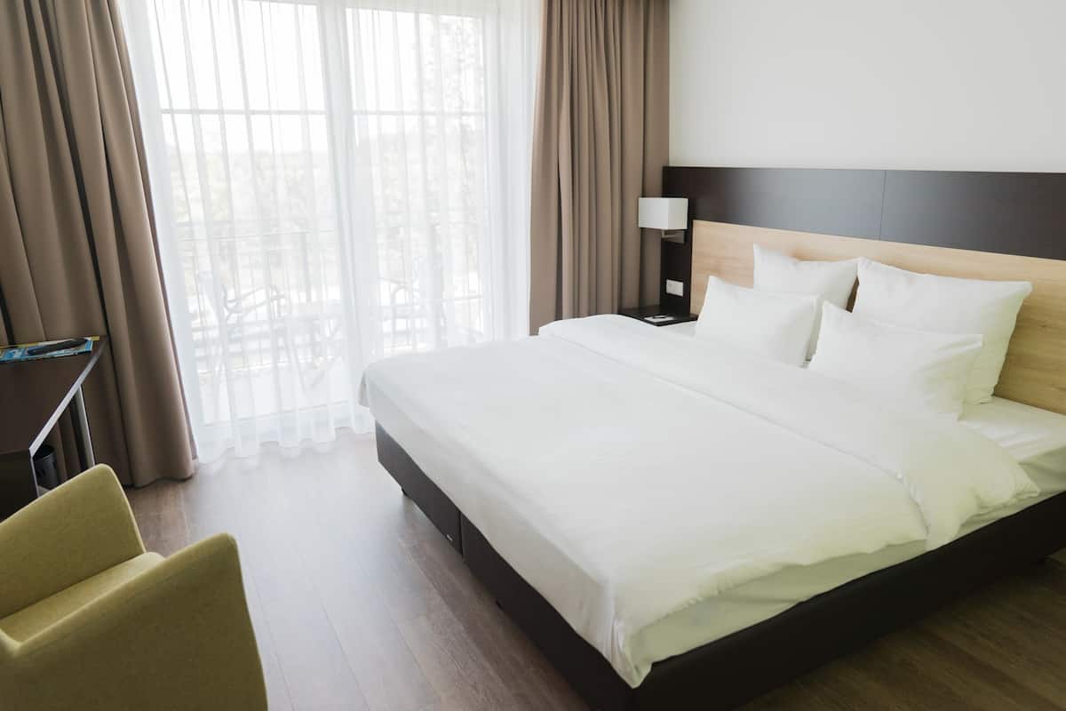 A hotel room with a large bed, a bedside table with a lamp, a chair, and a view of the outdoors through sheer curtains.
