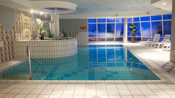 Una piscina interior con suelo de baldosas, zona central para nadar y área de descanso con sillas y una planta en maceta.