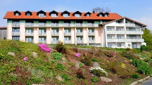 Un hotel de varios pisos con techo rojo y balcones. Se encuentra en una ladera con un jardín delantero.