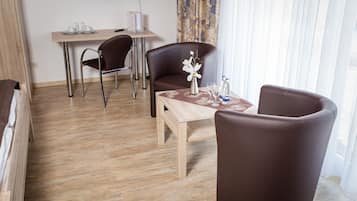 Habitación de hotel con piso de madera, una cama, un escritorio con dos sillas, una mesa con un florero y vasos, y vistas al exterior a través de cortinas translúcidas.