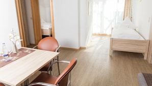 Habitación de hotel con cama de madera, escritorio de madera con silla, pequeña mesa con un florero y vasos, y una puerta que da a otra habitación.