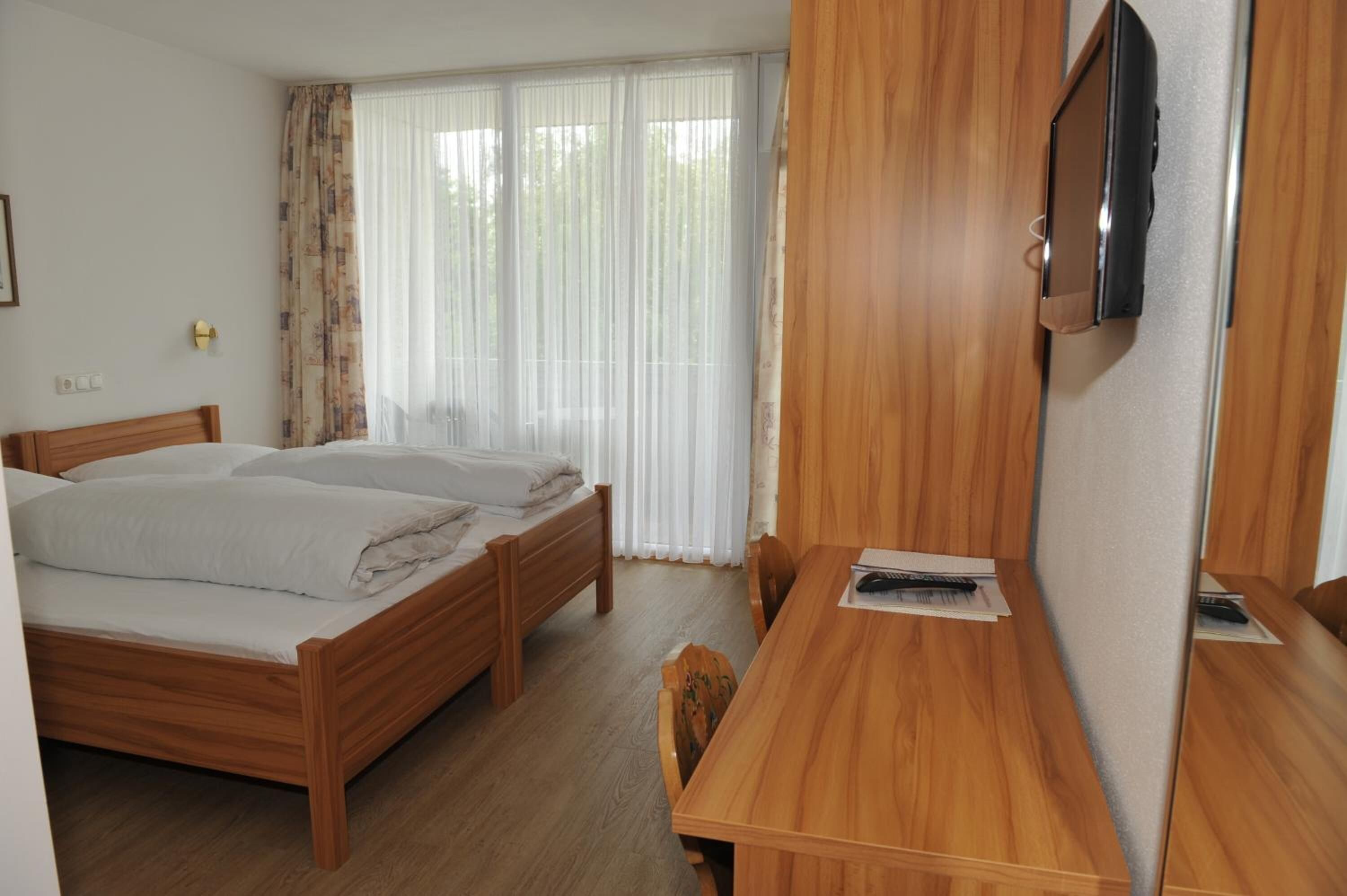 Une chambre d’hôtel avec deux lits en bois, un bureau en bois, une télévision et une fenêtre avec des rideaux légers.