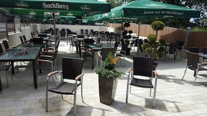 Un área de comedor al aire libre con mesas y sillas, sombrillas verdes y una planta en maceta.