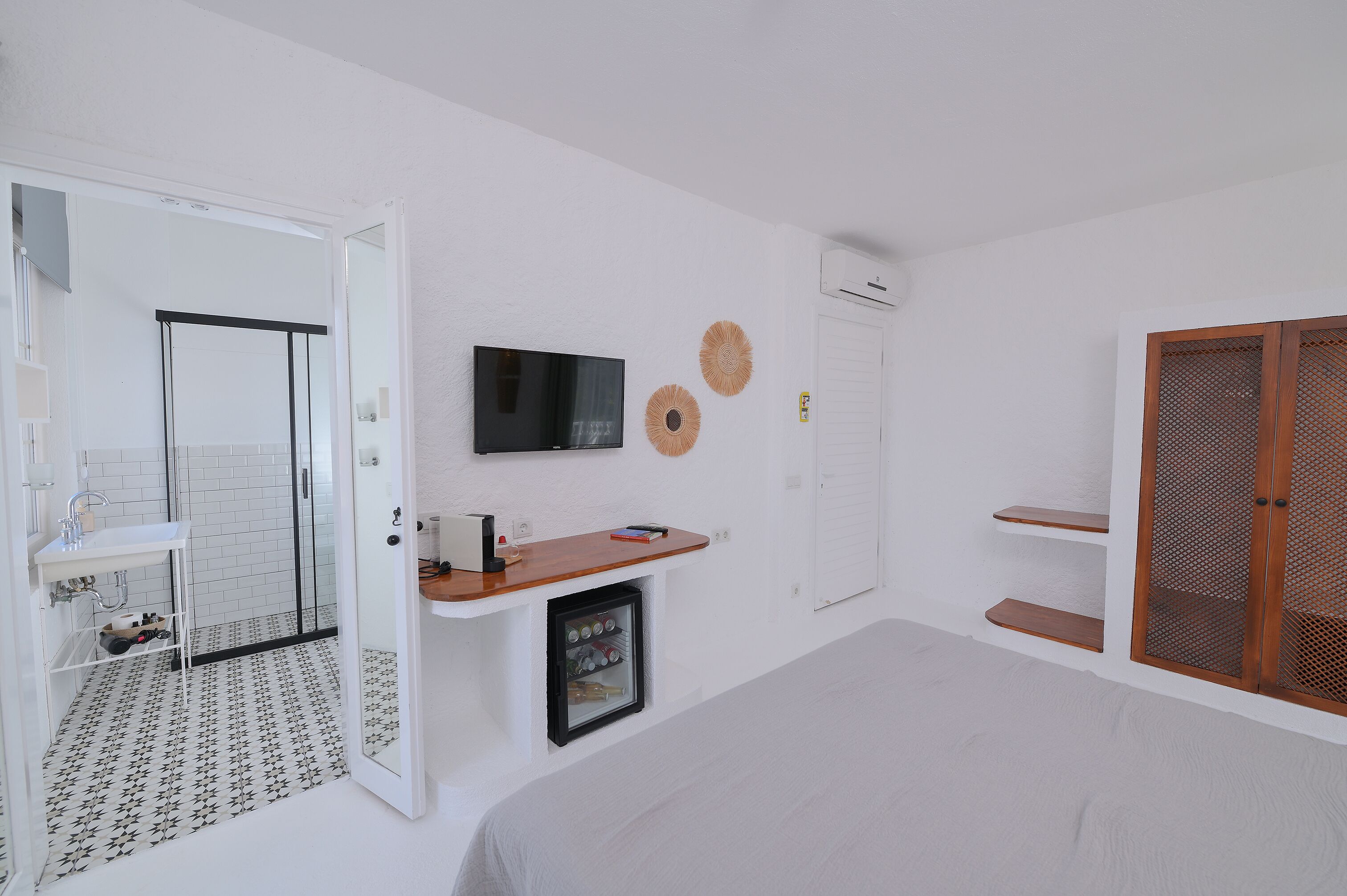 Un baño moderno con lavamanos, espejo y retrete blancos. Cuenta con zona de ducha con puerta de cristal, un refrigerador pequeño y un televisor montado en la pared.