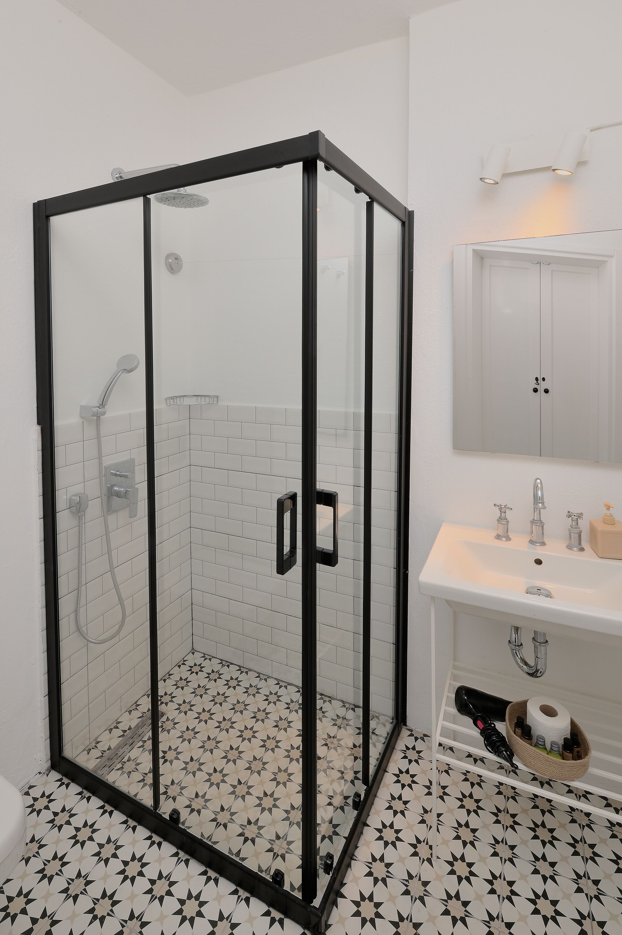 A modern bathroom with a glass-enclosed shower, white tiled walls, a white sink with a mirror above, and a patterned floor.