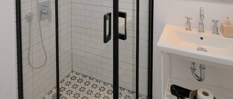 A modern bathroom with a glass-enclosed shower, white tiled walls, a white sink with a mirror above, and a patterned floor.