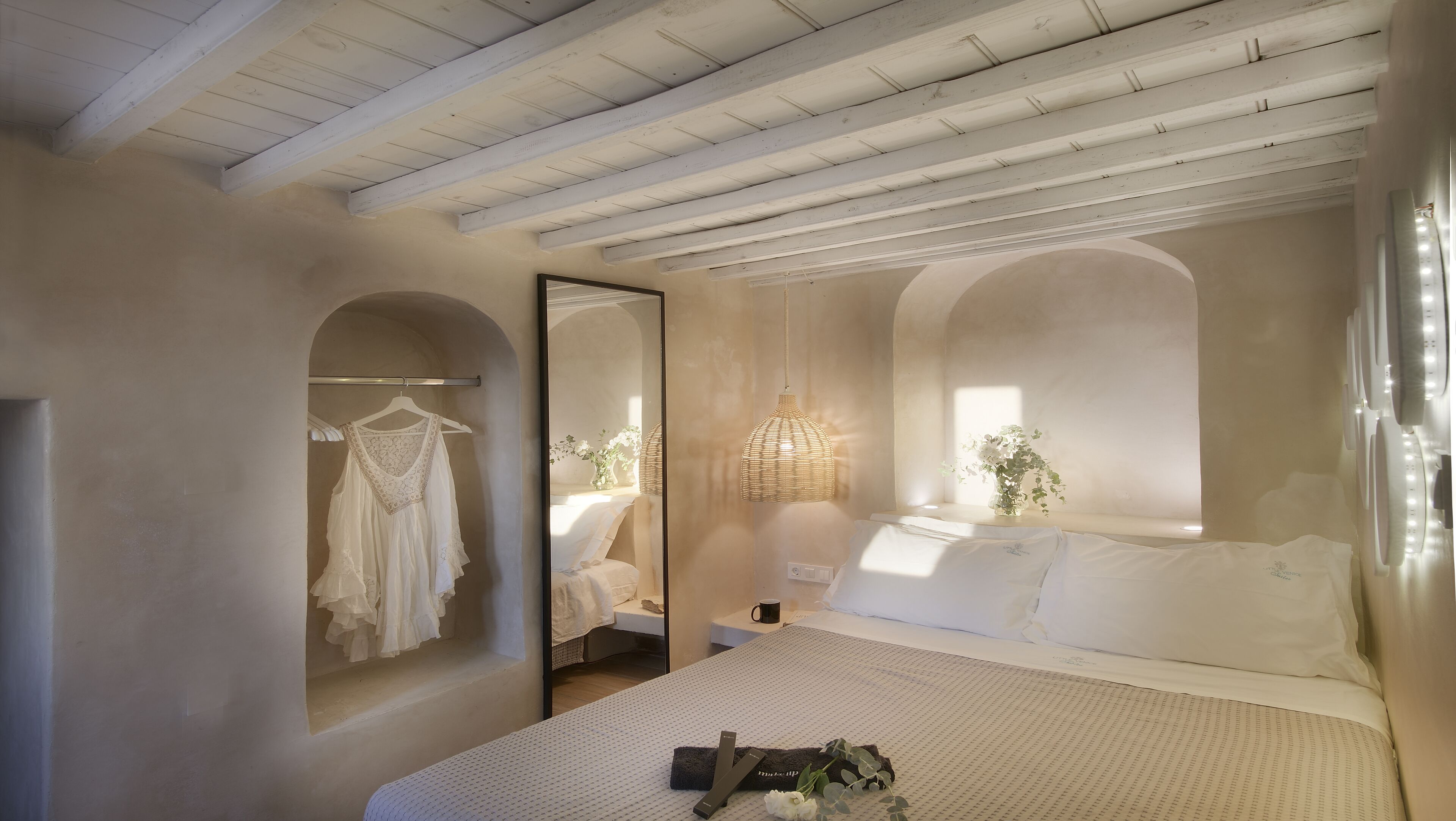 A bedroom with a large bed, a hanging dress rack, a mirror, and a bedside table with a lamp.