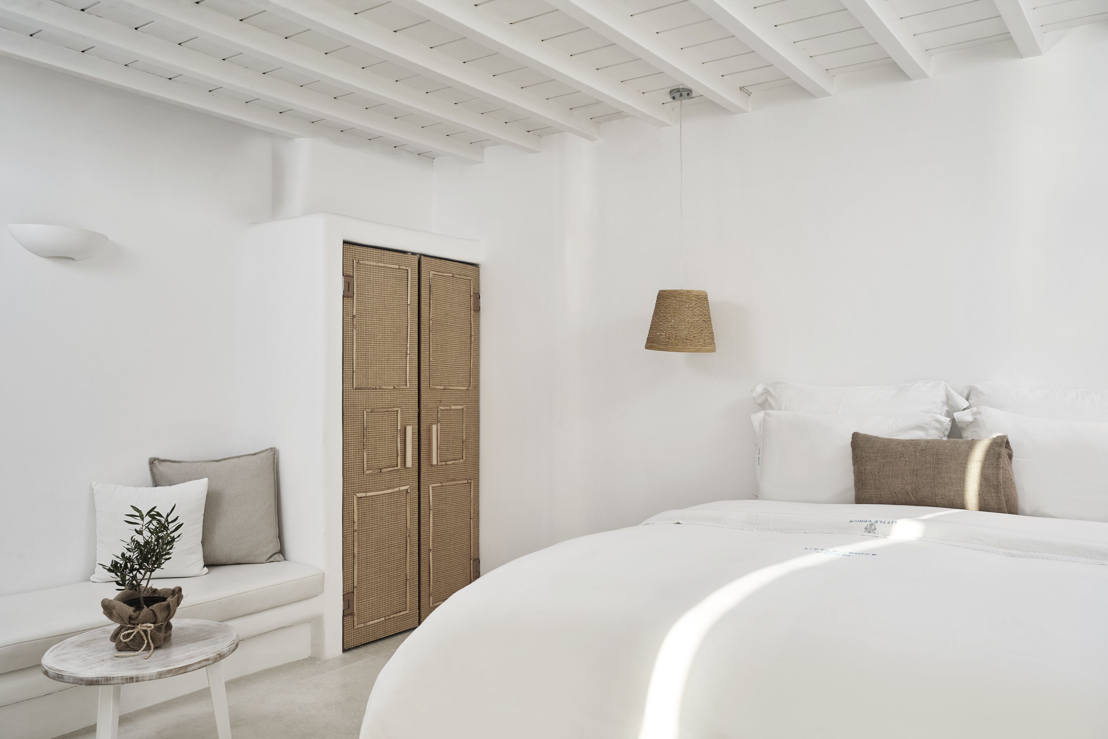 A modern, minimalist bedroom with a large bed, a small round table with a plant, a woven wardrobe, and a wall-mounted lamp.