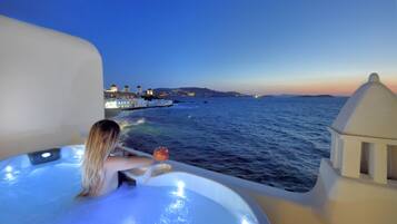 A person relaxing in a hot tub with a view of the sea and buildings at sunset.