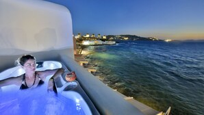 A person in a hot tub with a drink, overlooking a coastal area with buildings and a sea view.