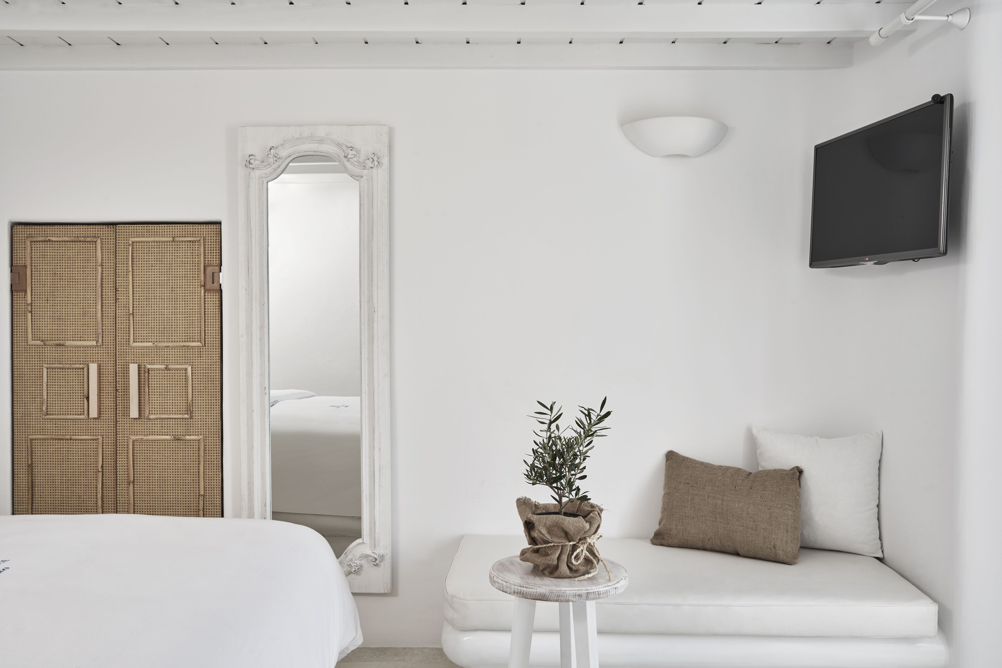 Quarto minimalista com uma cama, uma mesinha redonda com uma planta, um sofá com almofadas, um espelho grande e uma TV de tela plana.