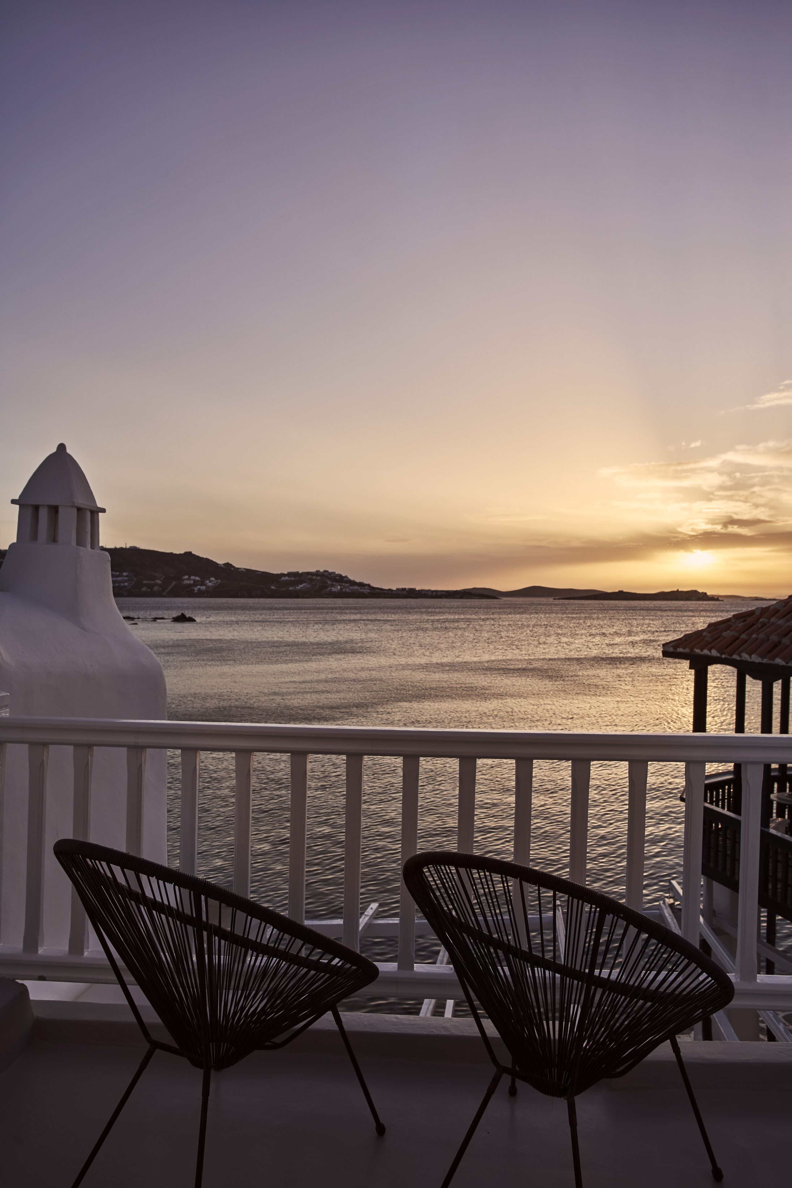 A balcony with two black chairs, a white railing, and a view of the sea at sunset.