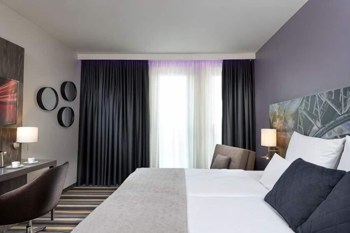 A hotel room with a bed, a desk, a chair, and a large window with curtains.