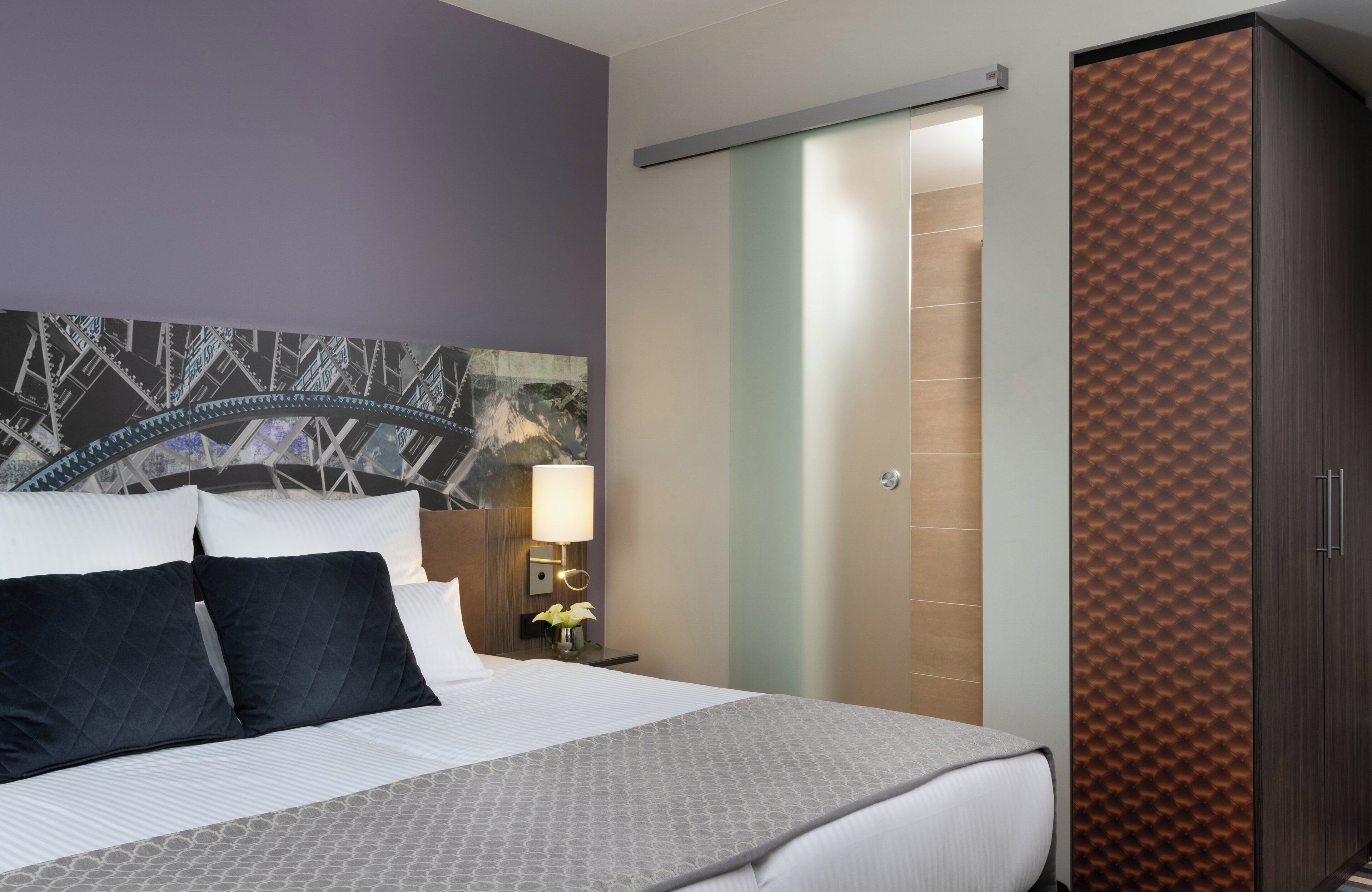 A hotel room with a bed, a nightstand, a lamp, a wardrobe, and a wall with a mural.