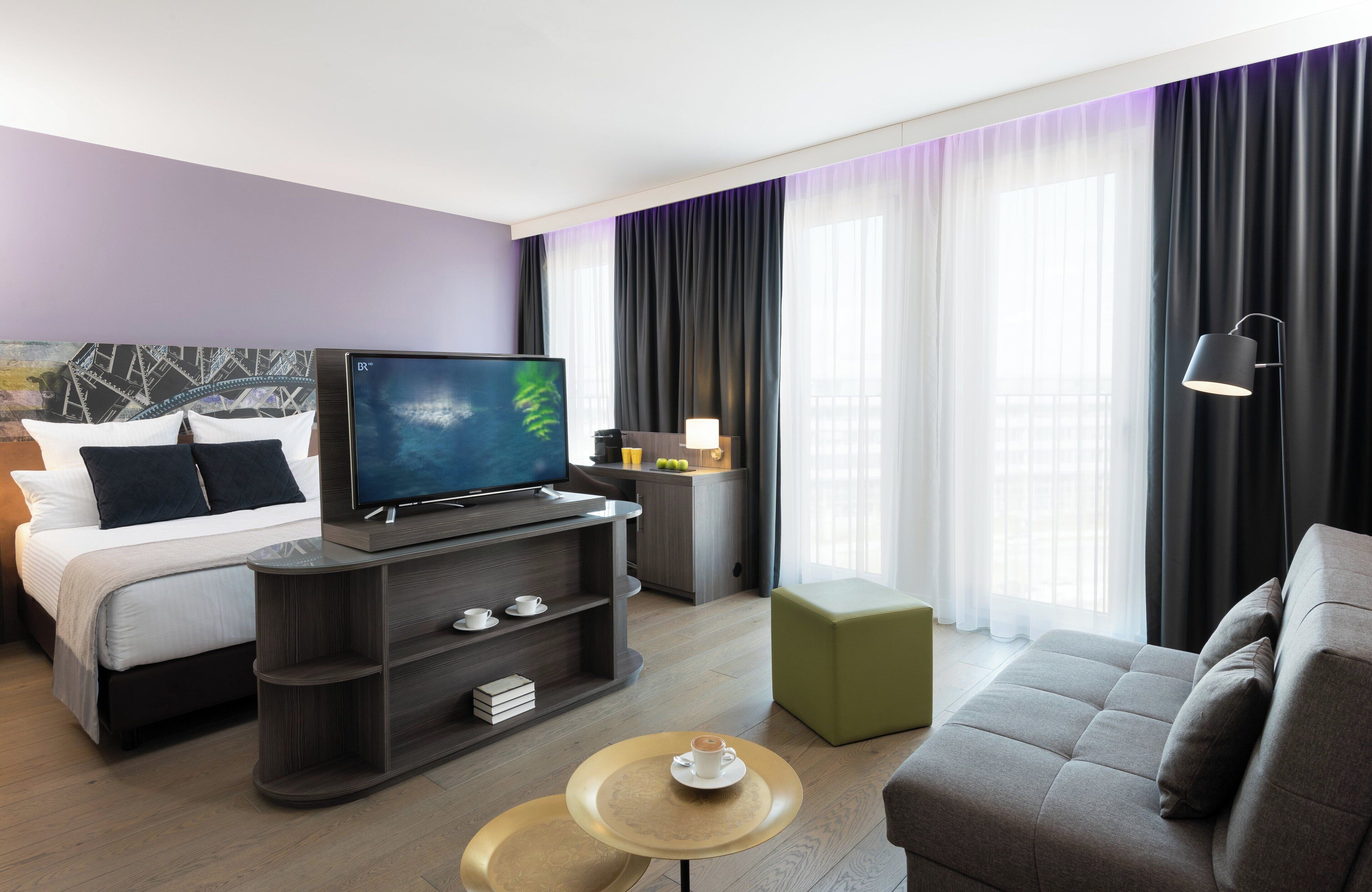 A modern hotel room with a bed, a TV, a sofa, a coffee table, and a small table with a lamp.
