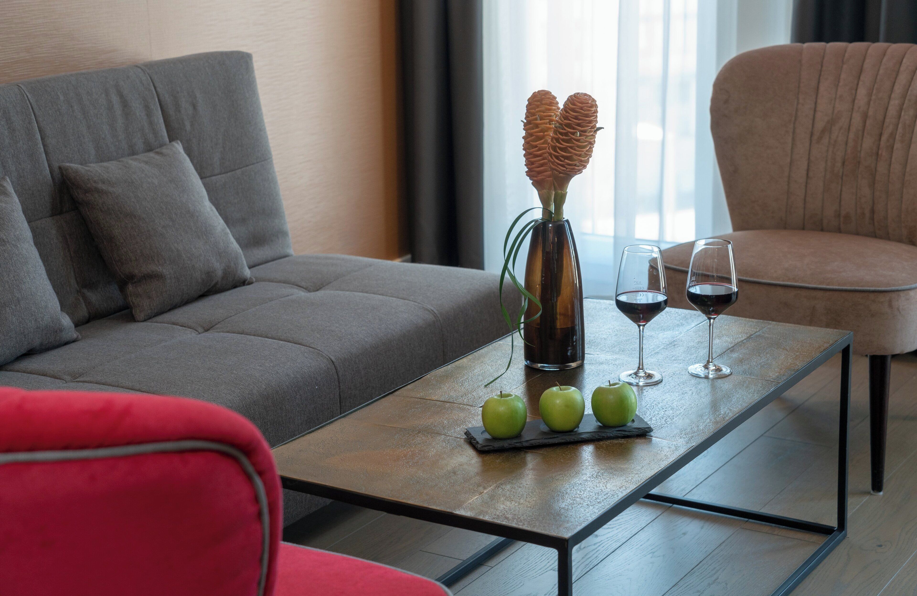 Ein modernes Wohnzimmer mit einem grauen Sofa, einem roten Sessel, einem Couchtisch mit grünen Äpfeln und Weingläsern sowie einer Vase mit dekorativen Zapfen.