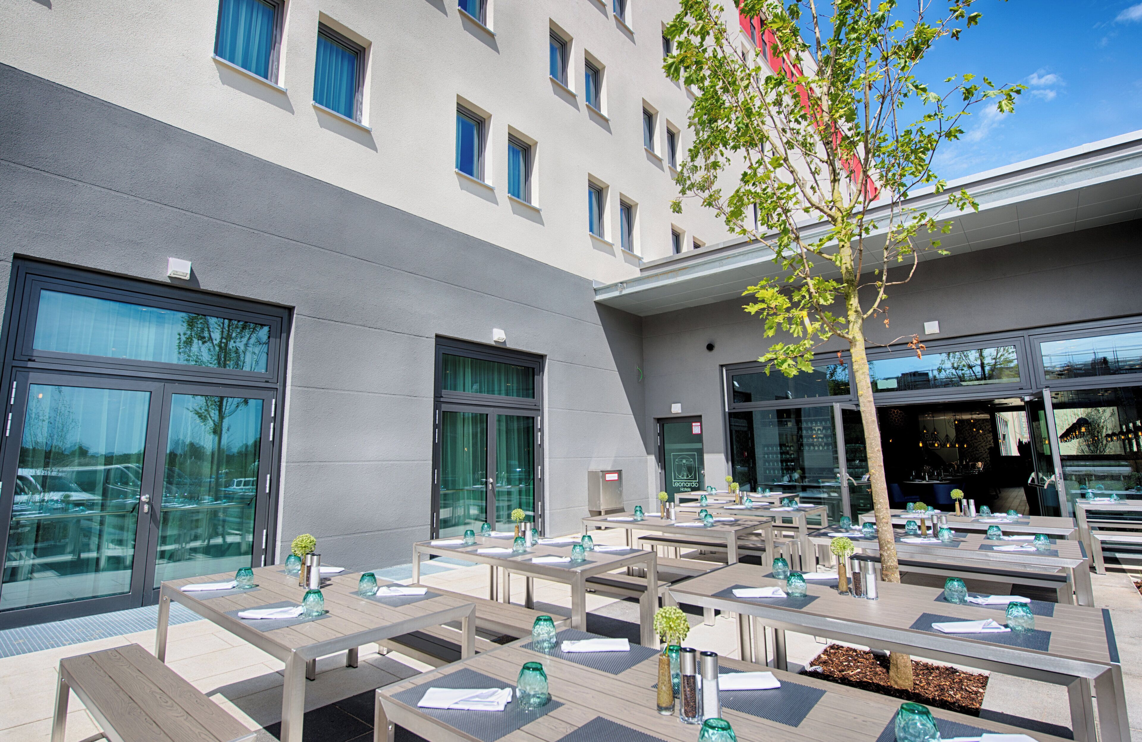 An outdoor dining area with multiple tables and chairs, a building with large windows, and a tree in the background.