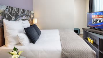 A hotel room with a bed, a TV displaying news, a painting on the wall, and a small table with a vase.