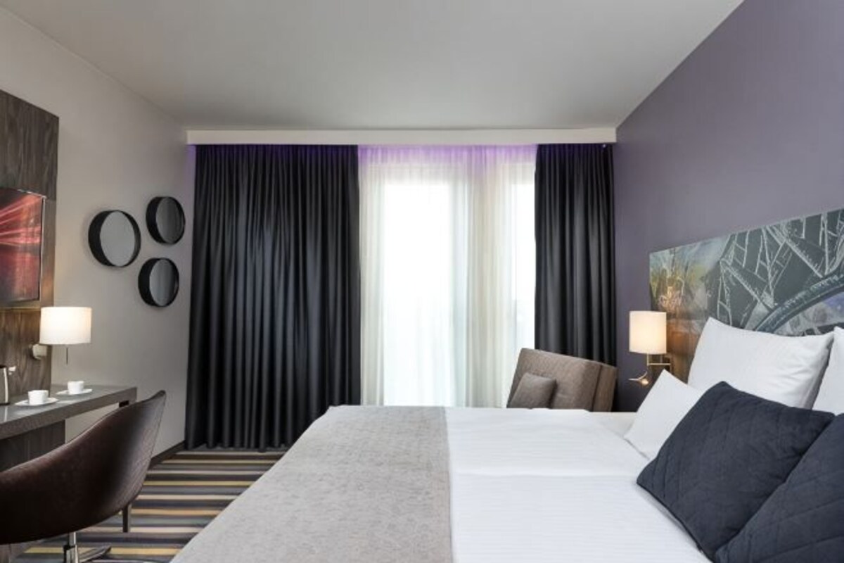 Una habitación de hotel con cama, escritorio, silla y un amplio ventanal con cortinas.