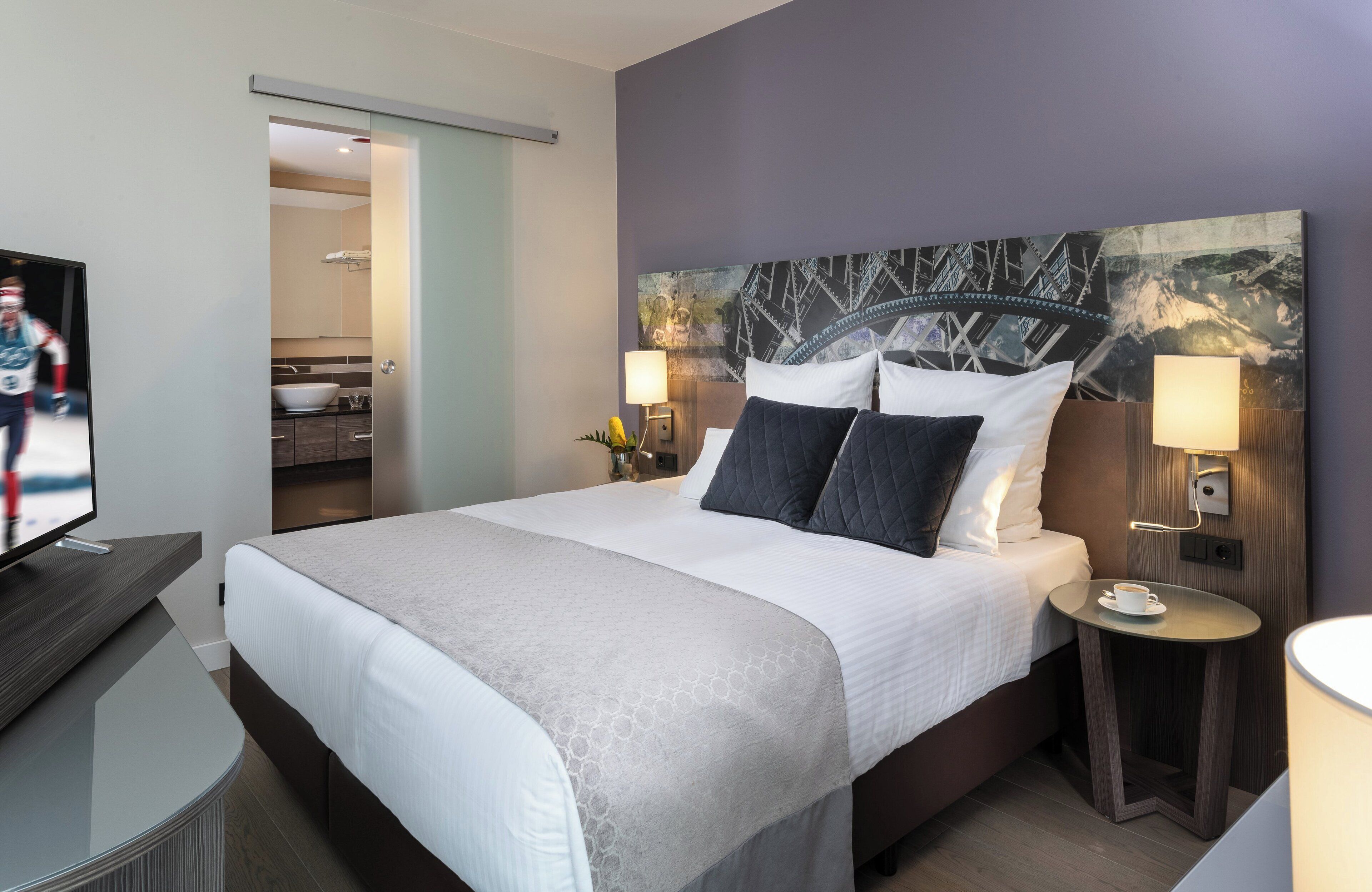 A modern hotel room with a large bed, a television, a small table with a cup, and a bathroom visible through an open door.