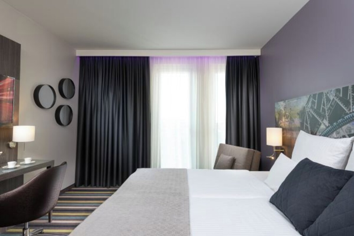 Habitación de hotel con una cama grande, un escritorio con silla, una ventana con cortinas y decoración en la pared.