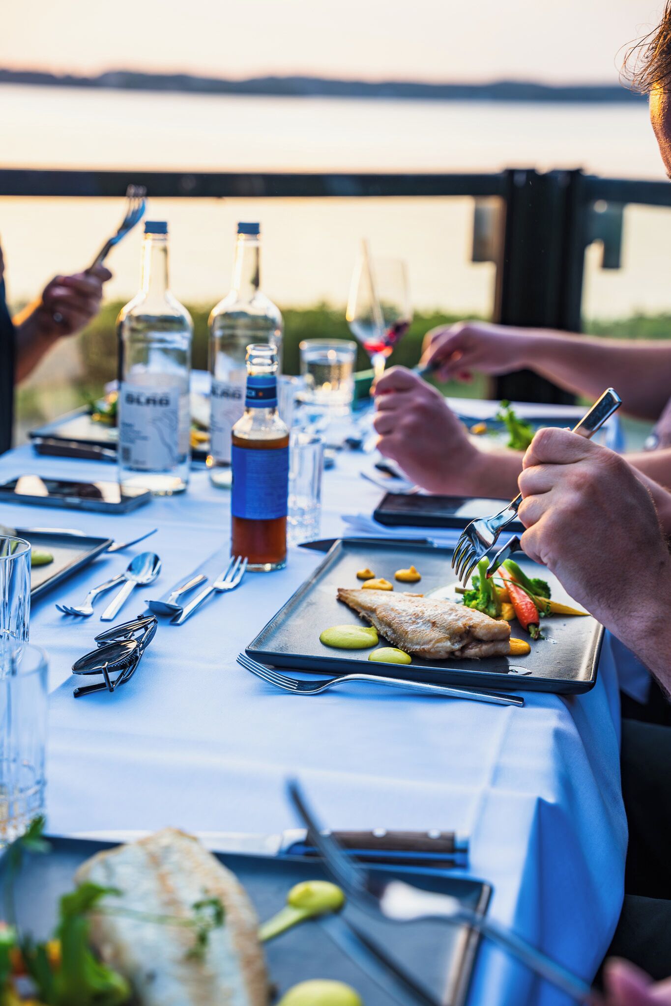 Una mesa de comedor dispuesta con platos, cubiertos y botellas, con vista al agua de fondo.