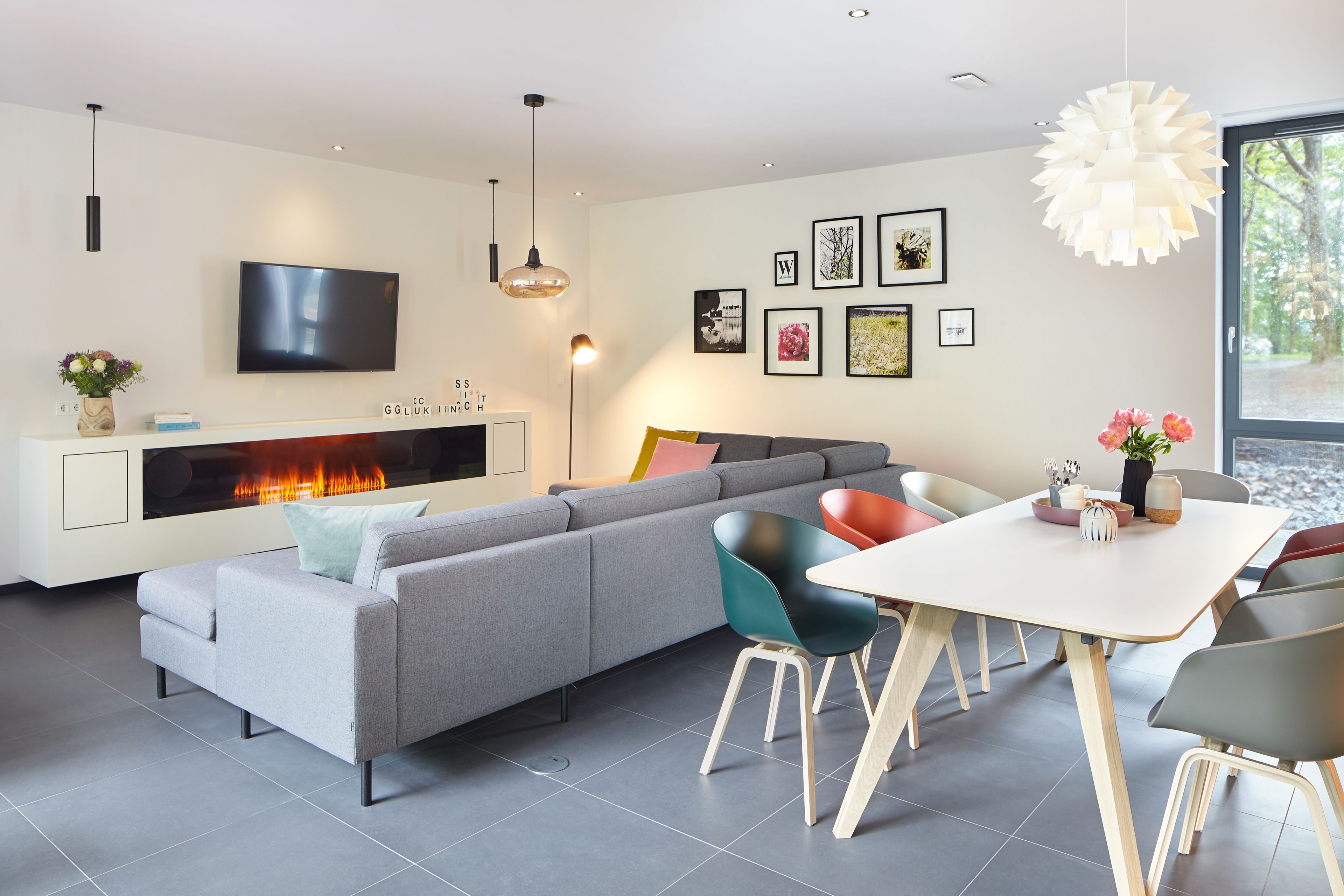 A modern living room with a grey sofa, teal and orange chairs, a white dining table, and a fireplace with a visible fire.