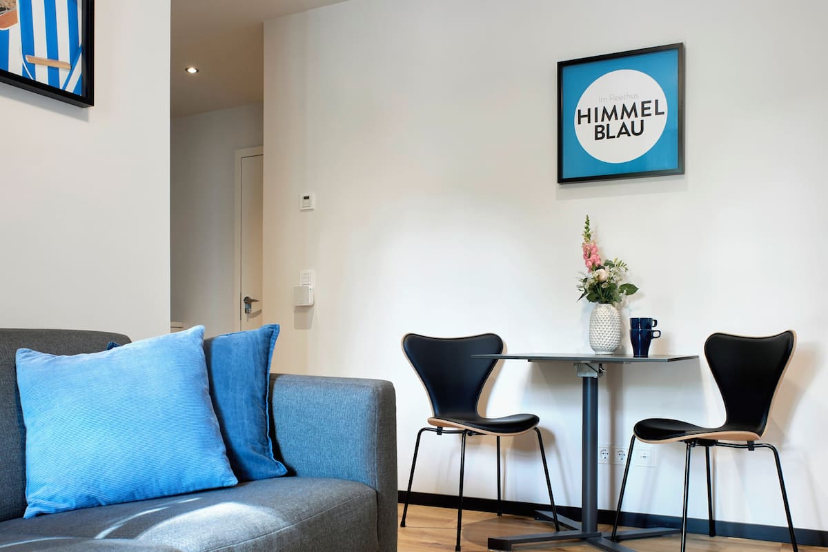 Ein modernes Wohnzimmer mit einem grauen Sofa, zwei schwarzen Sesseln, einem kleinen Tisch mit einer Vase und einer Tasse, sowie einem gerahmten Bild an der Wand.