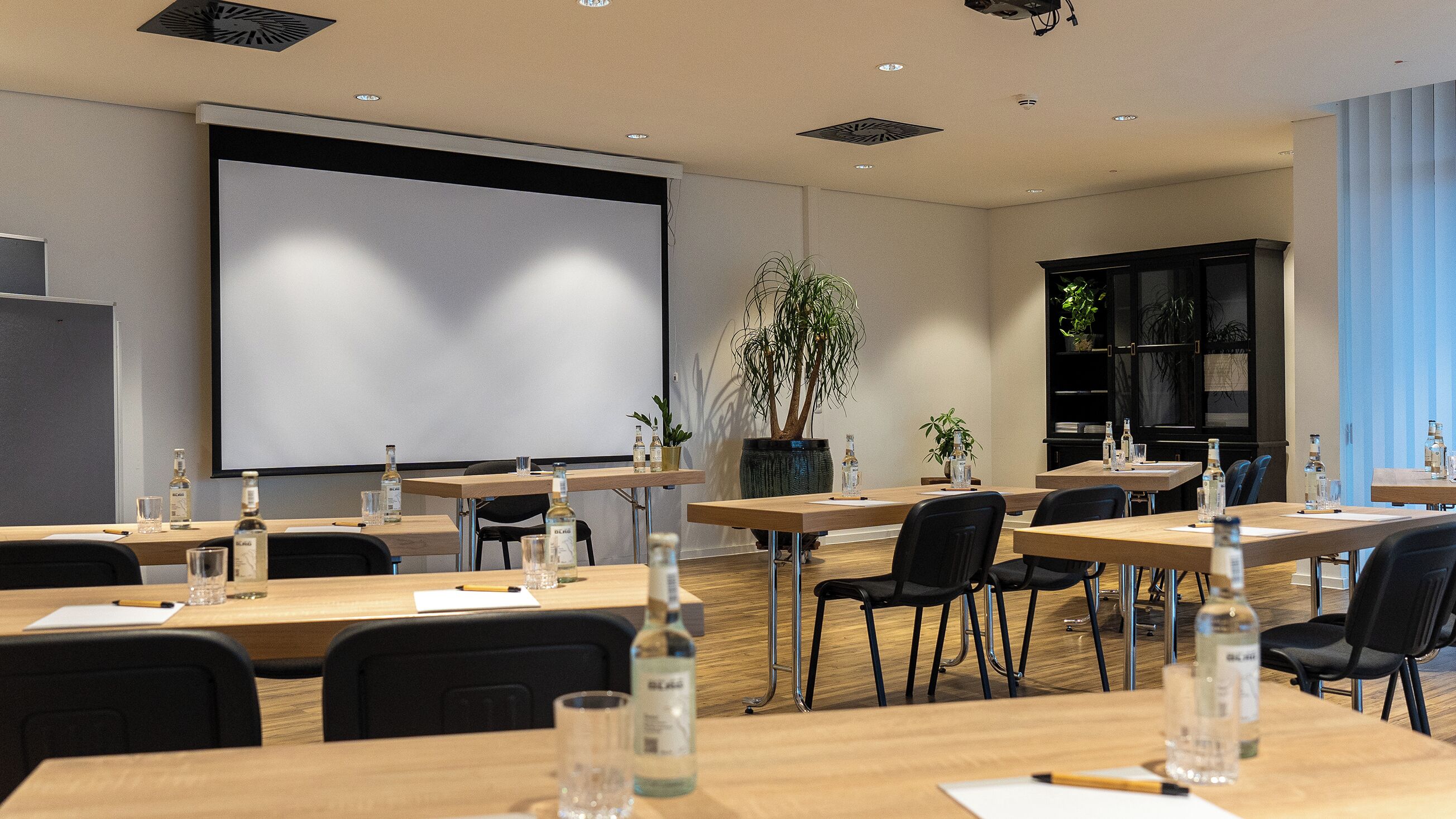 A conference room with tables, chairs, a projector screen, and a large potted plant.