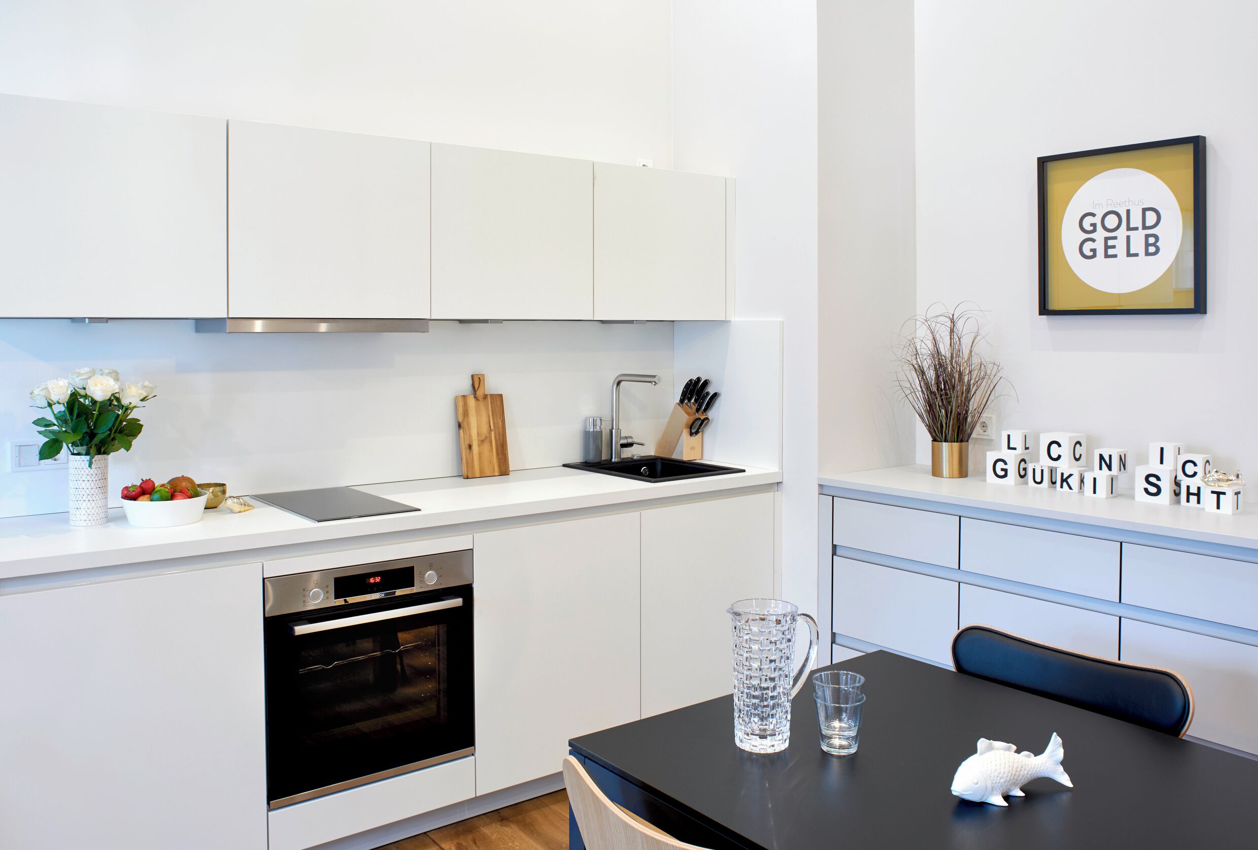 Une cuisine moderne avec des armoires blanches, un four en acier inoxydable, un évier et un coin repas comprenant une table et des chaises noires.