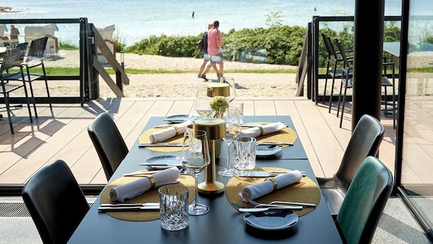Un espacio de comedor al aire libre con vista a la playa y al mar, mesas dispuestas con vajilla dorada y blanca, y sillas con tapicería negra y verde.