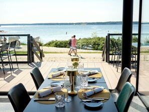 Un espacio de comedor al aire libre con vista a la playa y al mar, mesas dispuestas con vajilla dorada y blanca, y sillas con tapicería negra y verde.