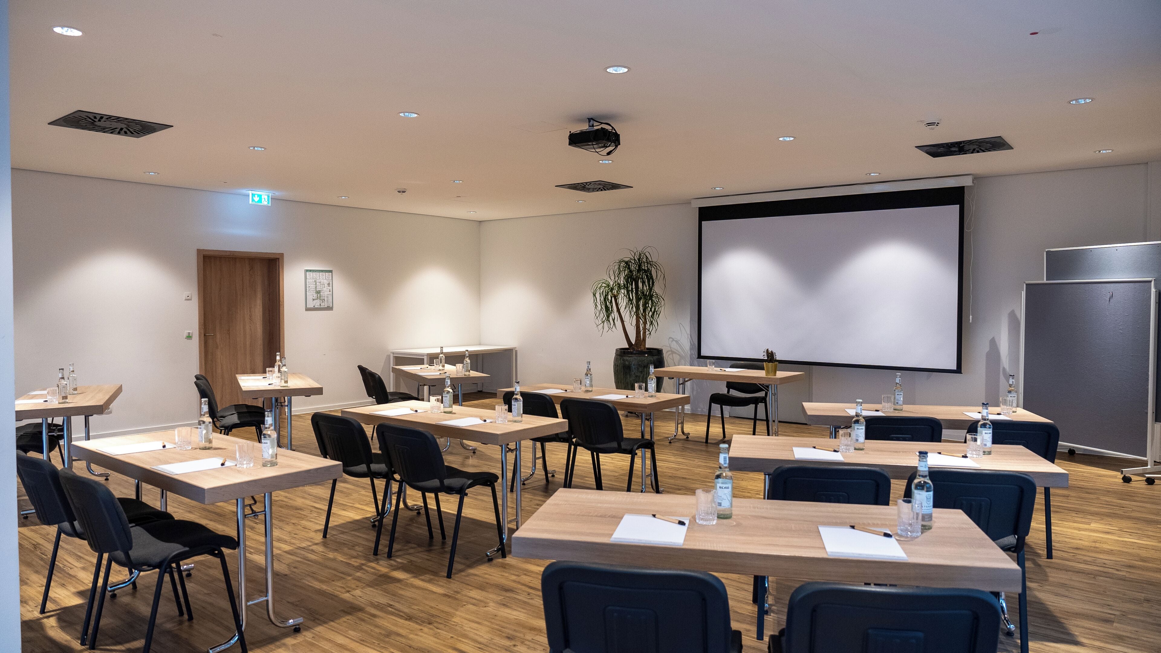 A conference room with tables, chairs, a projector screen, and a large plant.