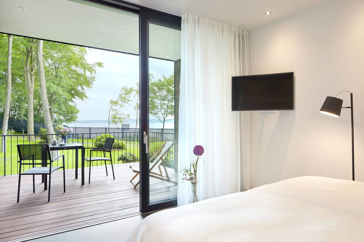 Ein Hotelzimmer mit einem Bett, einem Fernseher, einem Schreibtisch mit Stühlen und Blick auf eine Terrasse mit Tisch und Stühlen.