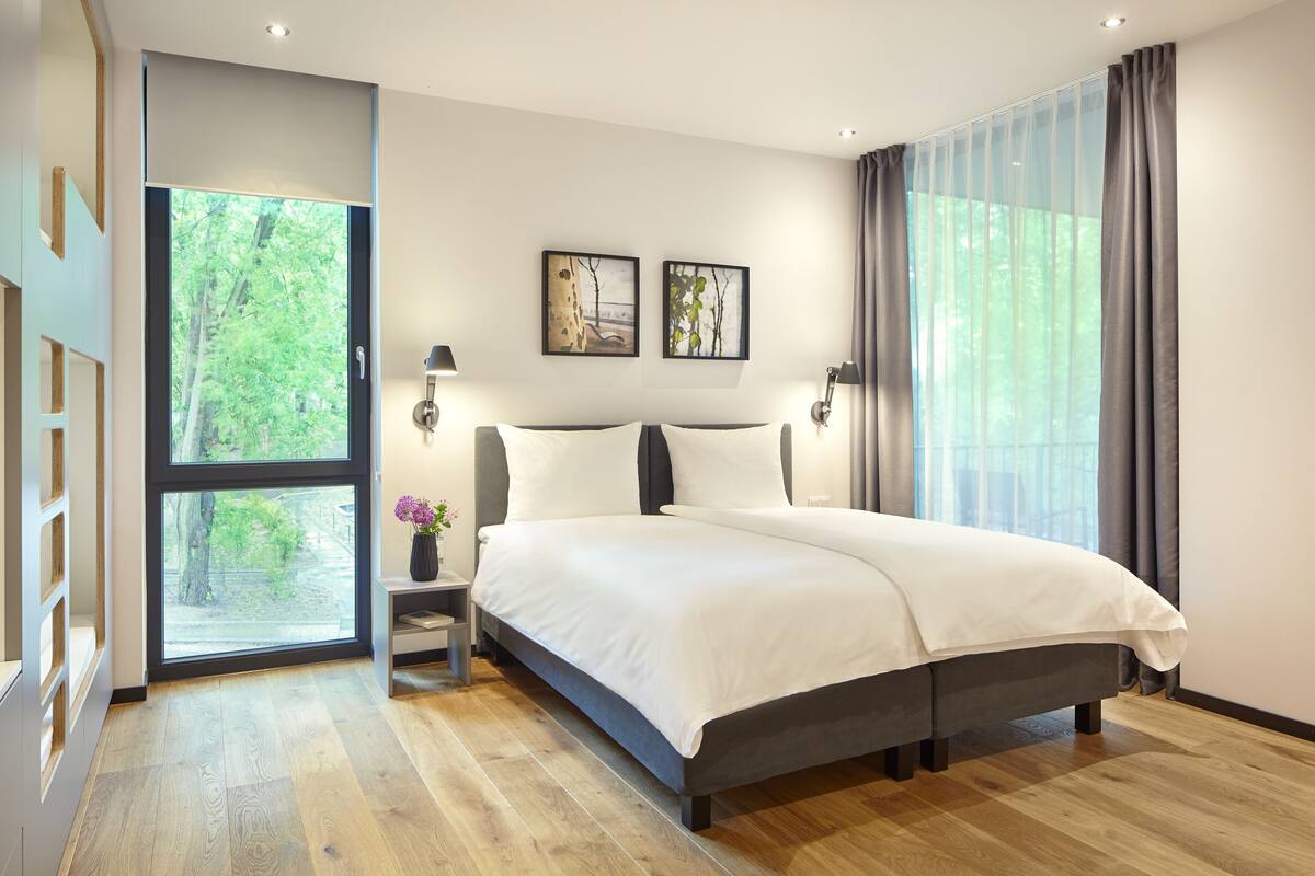 Ein modernes Schlafzimmer mit einem großen Bett, einem Nachttisch, einem Fenster mit Vorhängen und zwei gerahmten Bildern an der Wand.