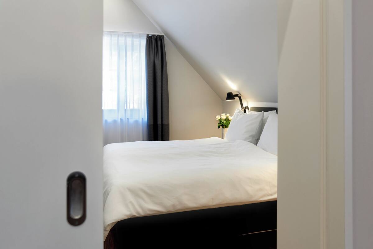 Ein Schlafzimmer mit einem Bett, einem Nachttisch, einer Lampe und einem Fenster mit Vorhängen.