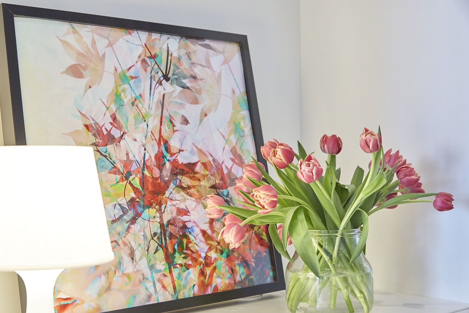Un cuadro abstracto enmarcado, un jarrón con tulipanes rosados y una lámpara sobre una mesa.