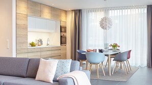 Una sala de estar moderna con un sofá gris, una mesa de comedor con sillas, una zona de cocina con microondas y un amplio ventanal con cortinas.