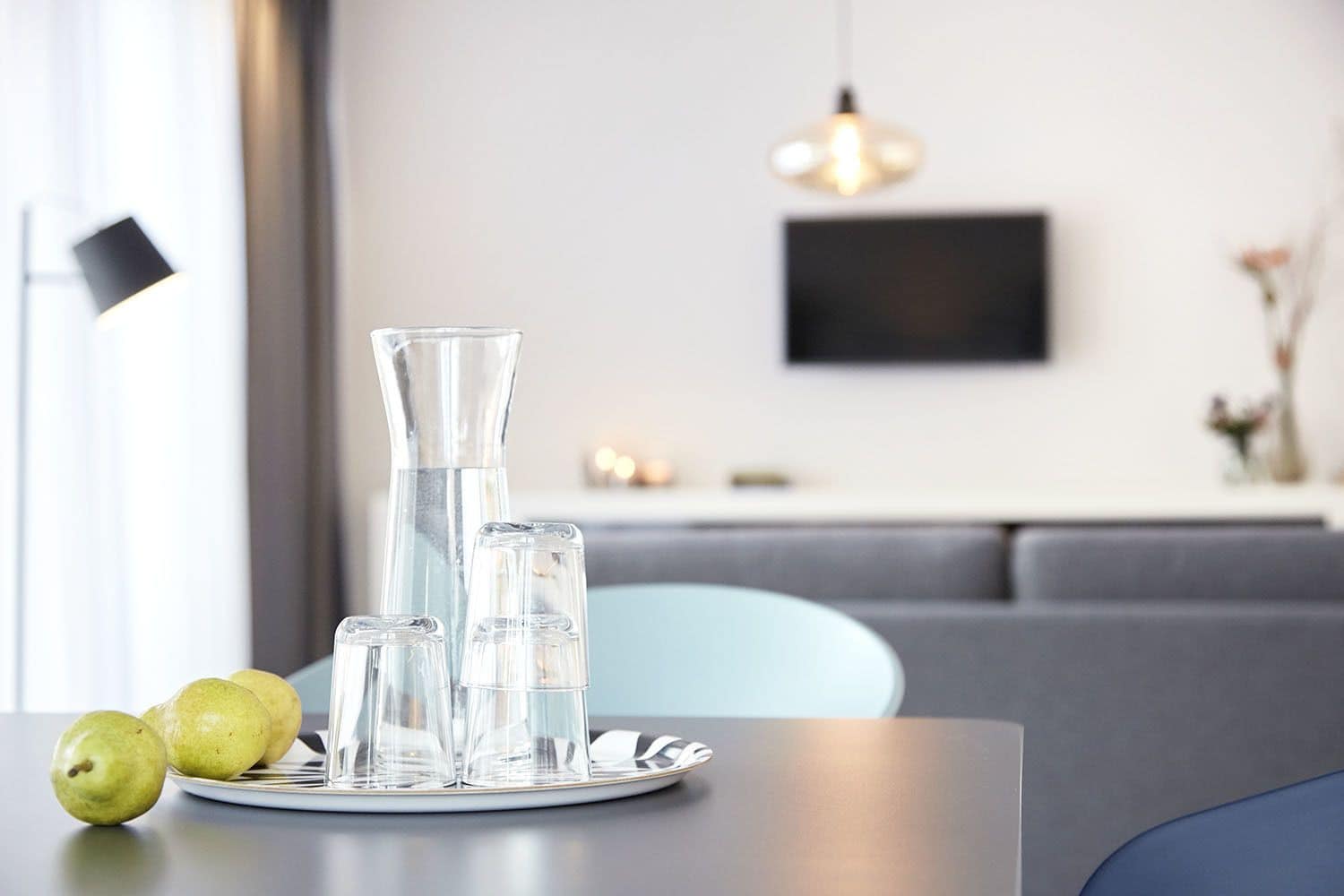 Una mesa de comedor con un plato de peras y cristalería transparente, un vaso alto y dos vasos más pequeños, ubicada en un moderno salón con un sofá gris y un televisor montado.