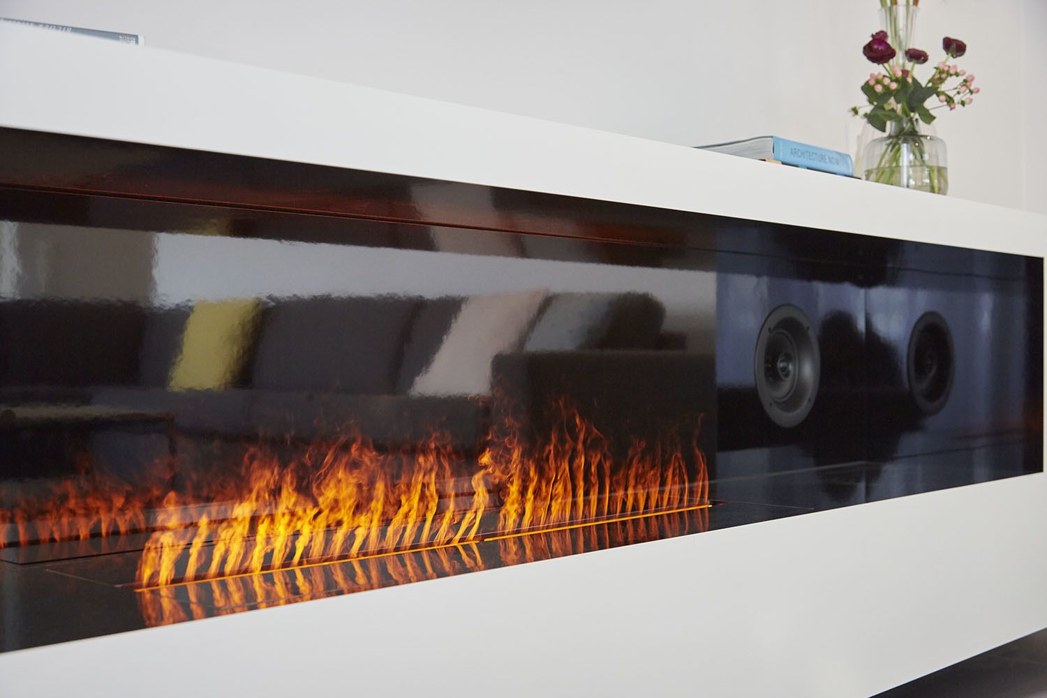A modern fireplace with a glass front, a sofa with cushions, and a vase with flowers on a white mantelpiece.