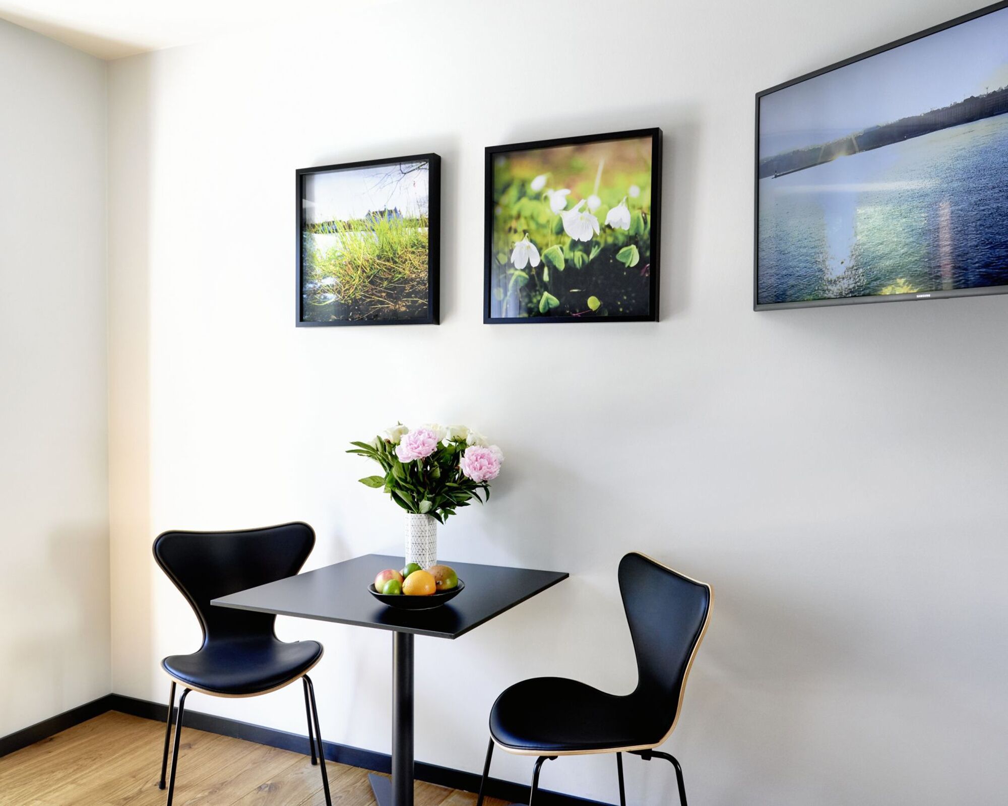 A room with a table, two chairs, a vase with flowers, and three framed pictures on the wall.