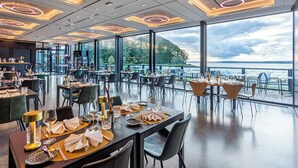 Un restaurante con mesas dispuestas para comer, amplias ventanas que ofrecen vistas a un cuerpo de agua y vegetación, y modernas lámparas de techo circulares.