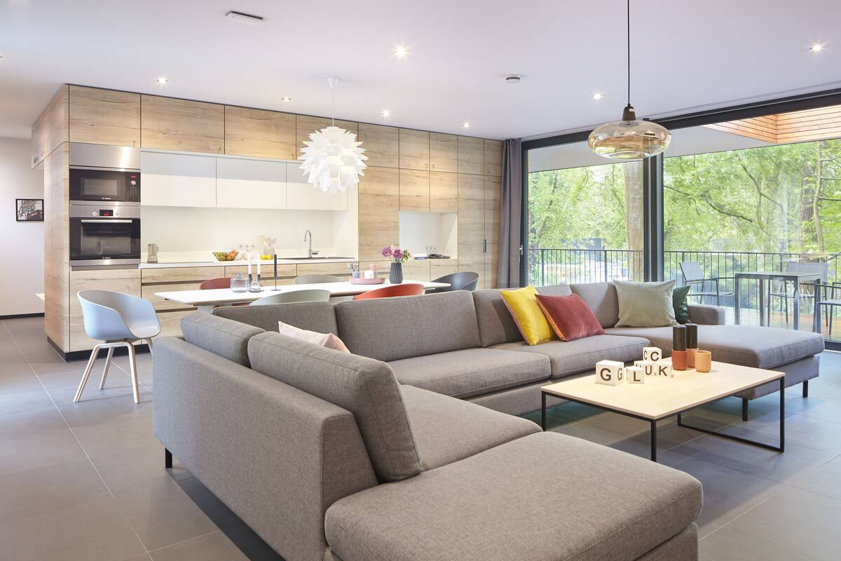 Ein modernes Wohnzimmer mit einem Ecksofa, einem Couchtisch und einem Essbereich, im Hintergrund ist eine Küche zu sehen.