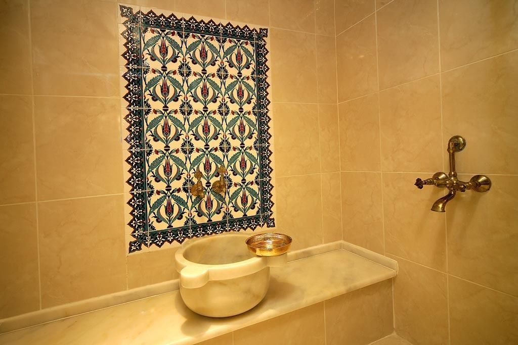 Un baño con pared de azulejos color beige, un panel decorativo de azulejos y un lavamanos blanco con un recipiente, además de un grifo de latón.