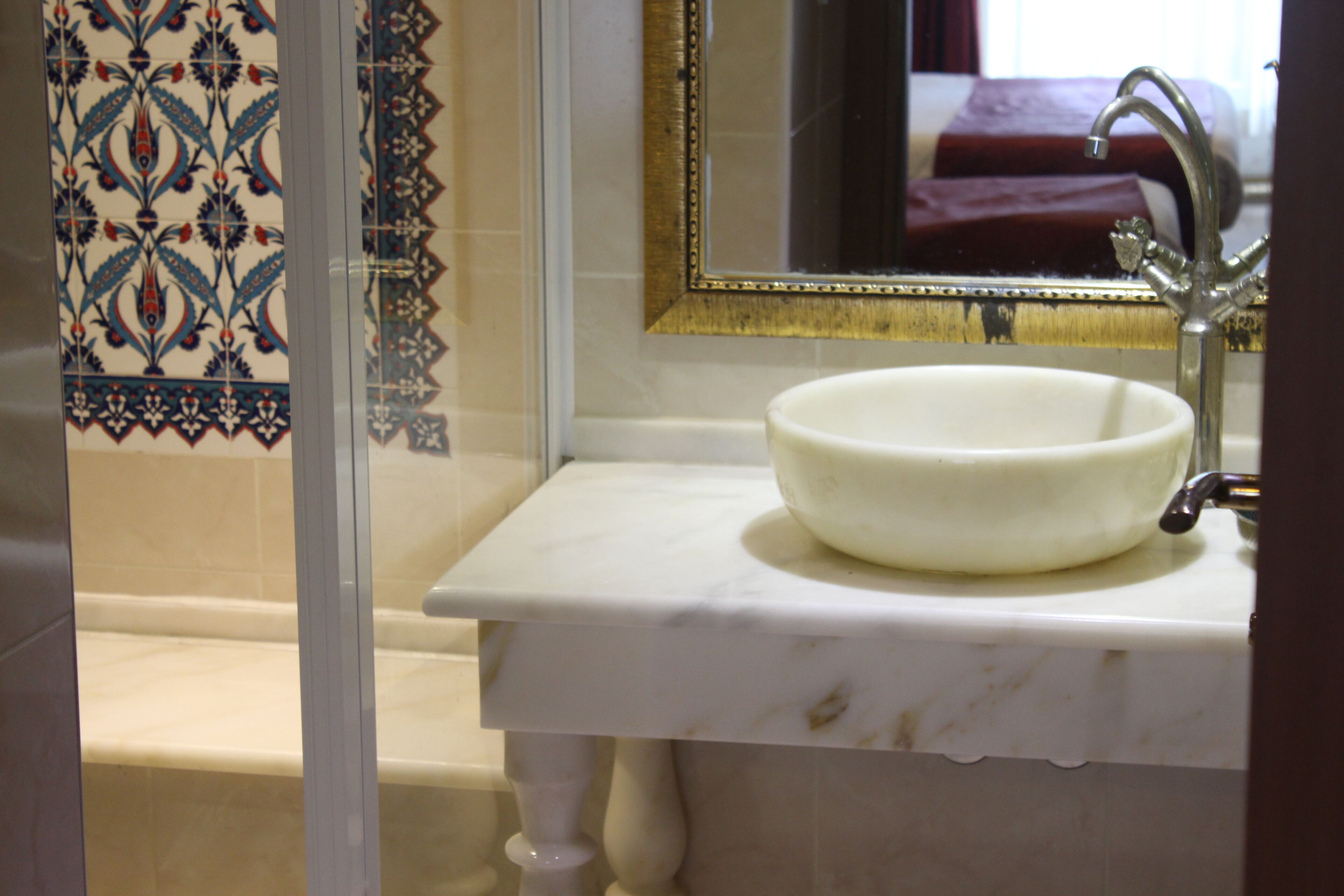 Un bany amb un lavabo de marbre blanc, un mirall amb marc daurat i rajoles amb dibuixos.