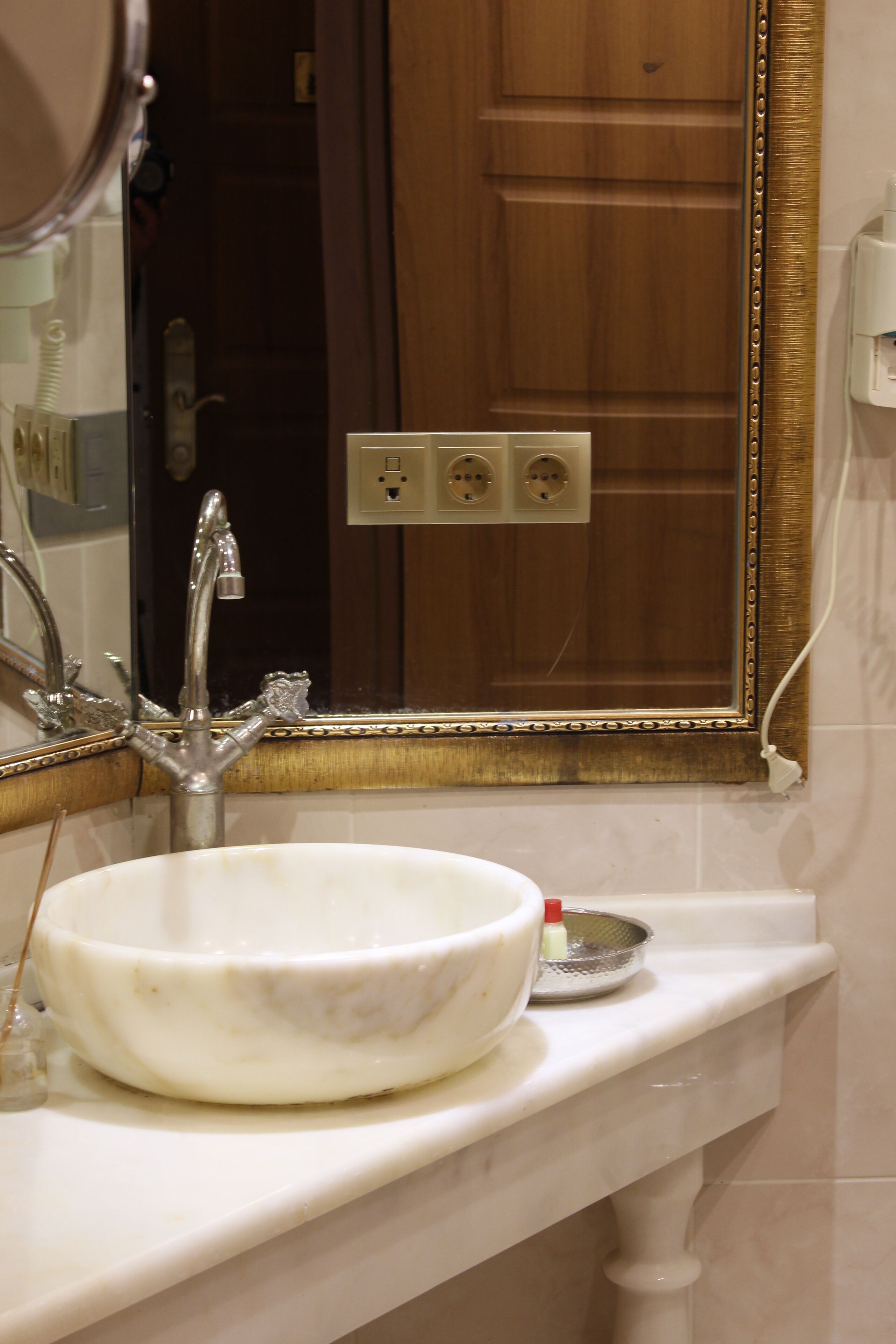Un baño con un lavamanos grande de color blanco, un espejo con marco dorado y una puerta de madera al fondo.