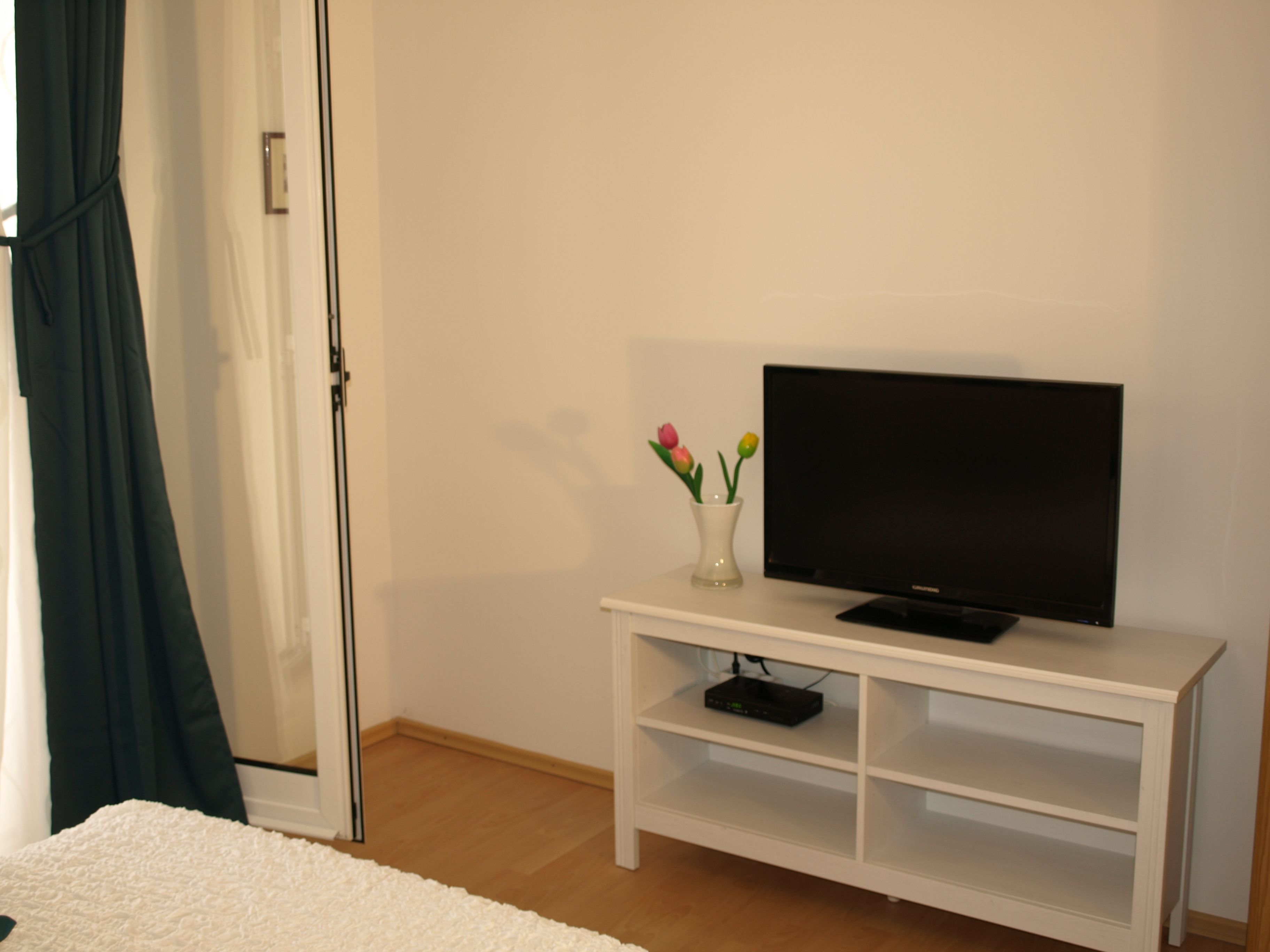 Une pièce avec un meuble blanc pour la télévision, un téléviseur à écran plat, un petit vase blanc contenant des fleurs et un tapis blanc.