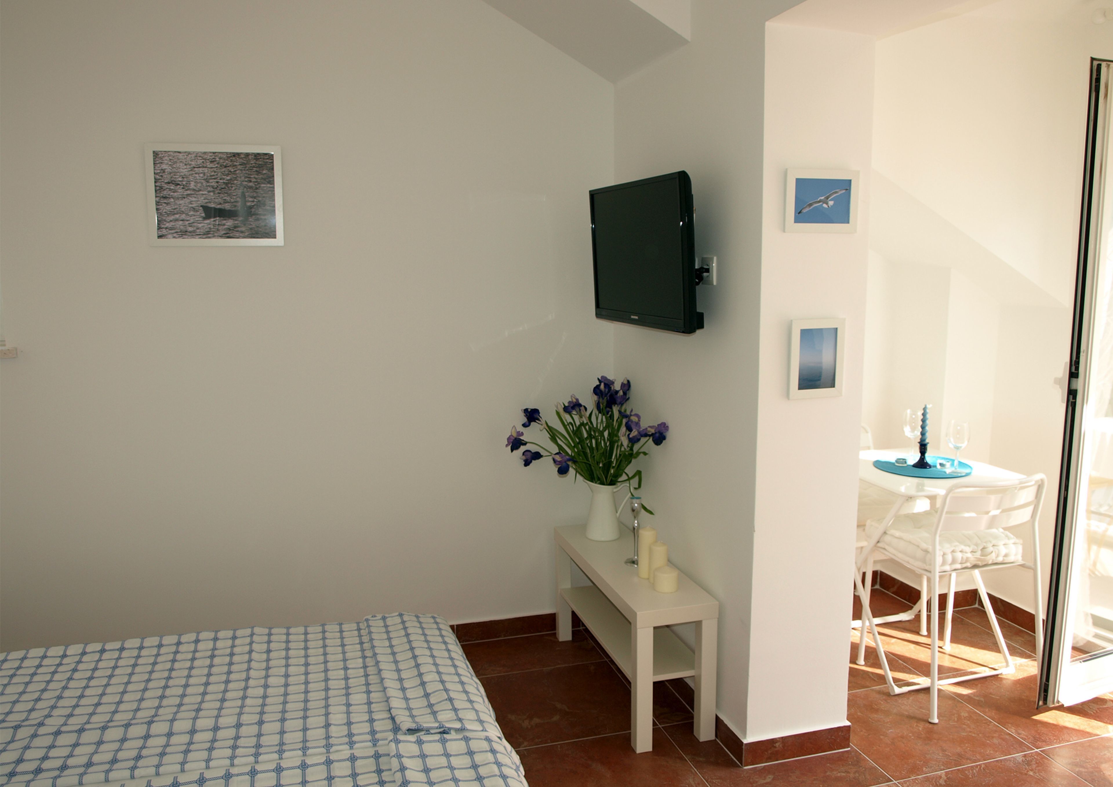 A bedroom with a bed, a small white table with a vase of flowers, a TV mounted on the wall, and a dining area with a table and chairs.