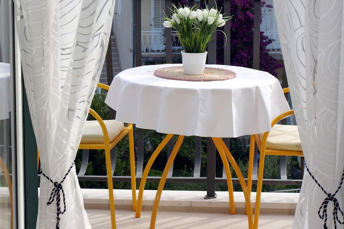 Un balcon avec une table ronde, des chaises jaunes, une plante en pot et des rideaux blancs.