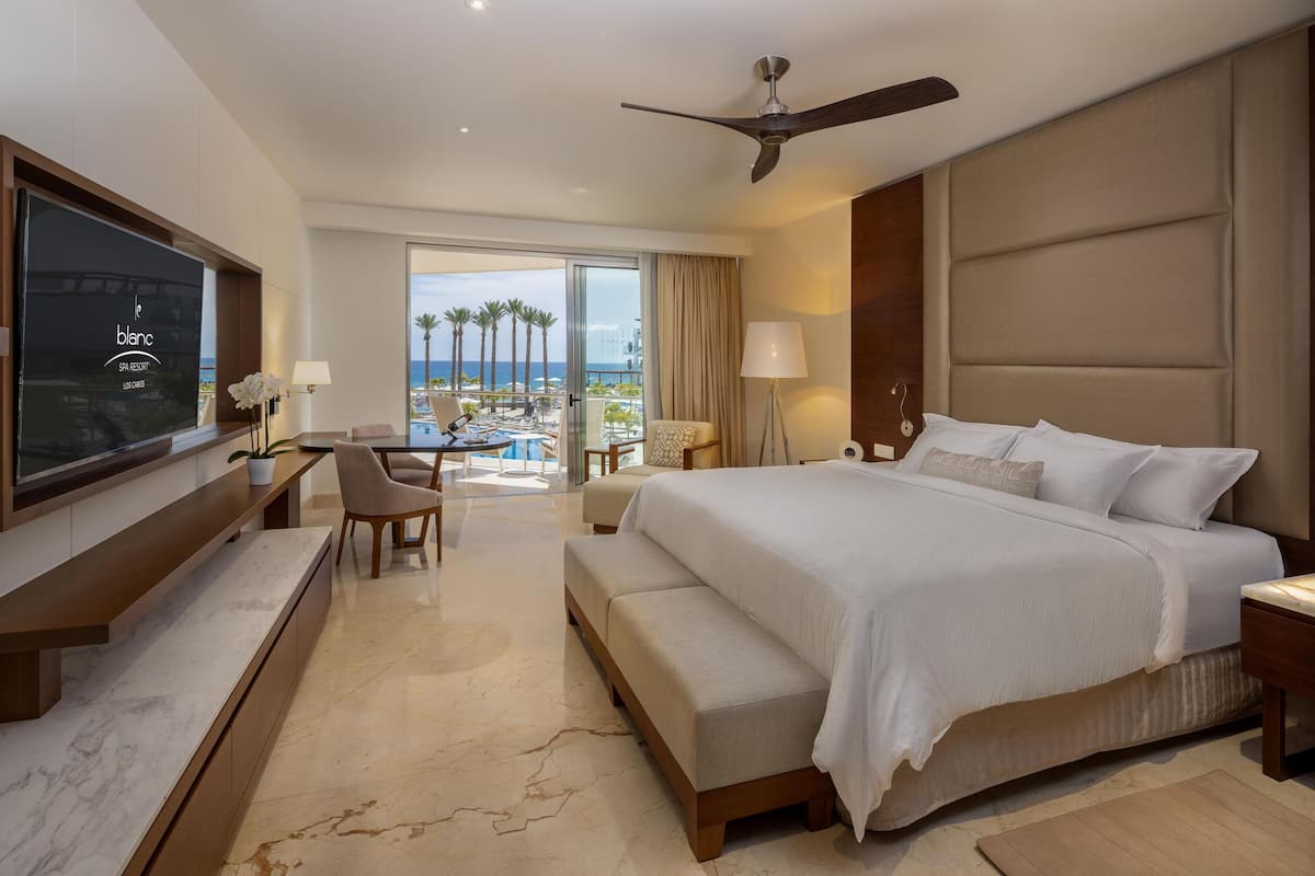 Une chambre d’hôtel avec un grand lit, un bureau avec une télévision, un balcon avec vue sur l’océan et un ventilateur de plafond.